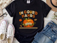 An black short-sleeve t-shirt with a colorful graphic on the front. The graphic features the text 'IN GOUD HANDS' in a stylized orange and brown font above a smiling orange pumpkin with a toothy grin and autumn-themed details.