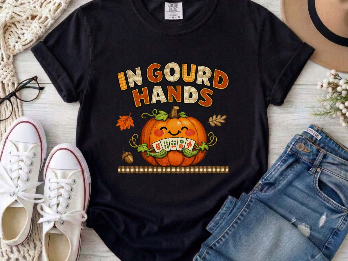 An black short-sleeve t-shirt with a colorful graphic on the front. The graphic features the text 'IN GOUD HANDS' in a stylized orange and brown font above a smiling orange pumpkin with a toothy grin and autumn-themed details.