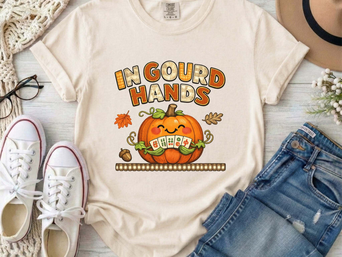 An ivory short-sleeve t-shirt with a colorful graphic on the front. The graphic features the text 'IN GOUD HANDS' in a stylized orange and brown font above a smiling orange pumpkin with a toothy grin and autumn-themed details.