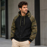 A man wearing a hybrid raglan hoodie with a solid black body and green-brown camouflage sleeves, paired with beige pants, standing in front of a modern concrete building.