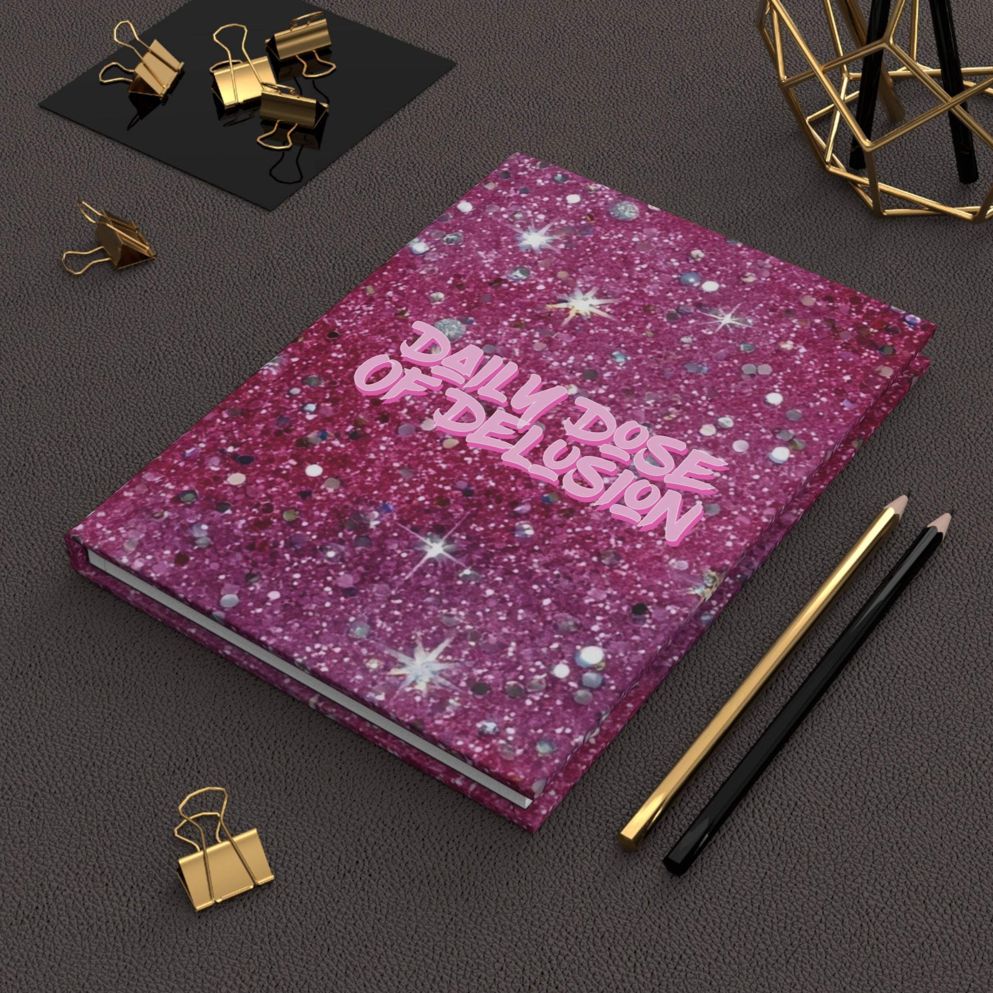 Hardcover journal with glittery pink cover titled Daily Dose of Delusion on black desk with gold and black pencils and gold binder clips