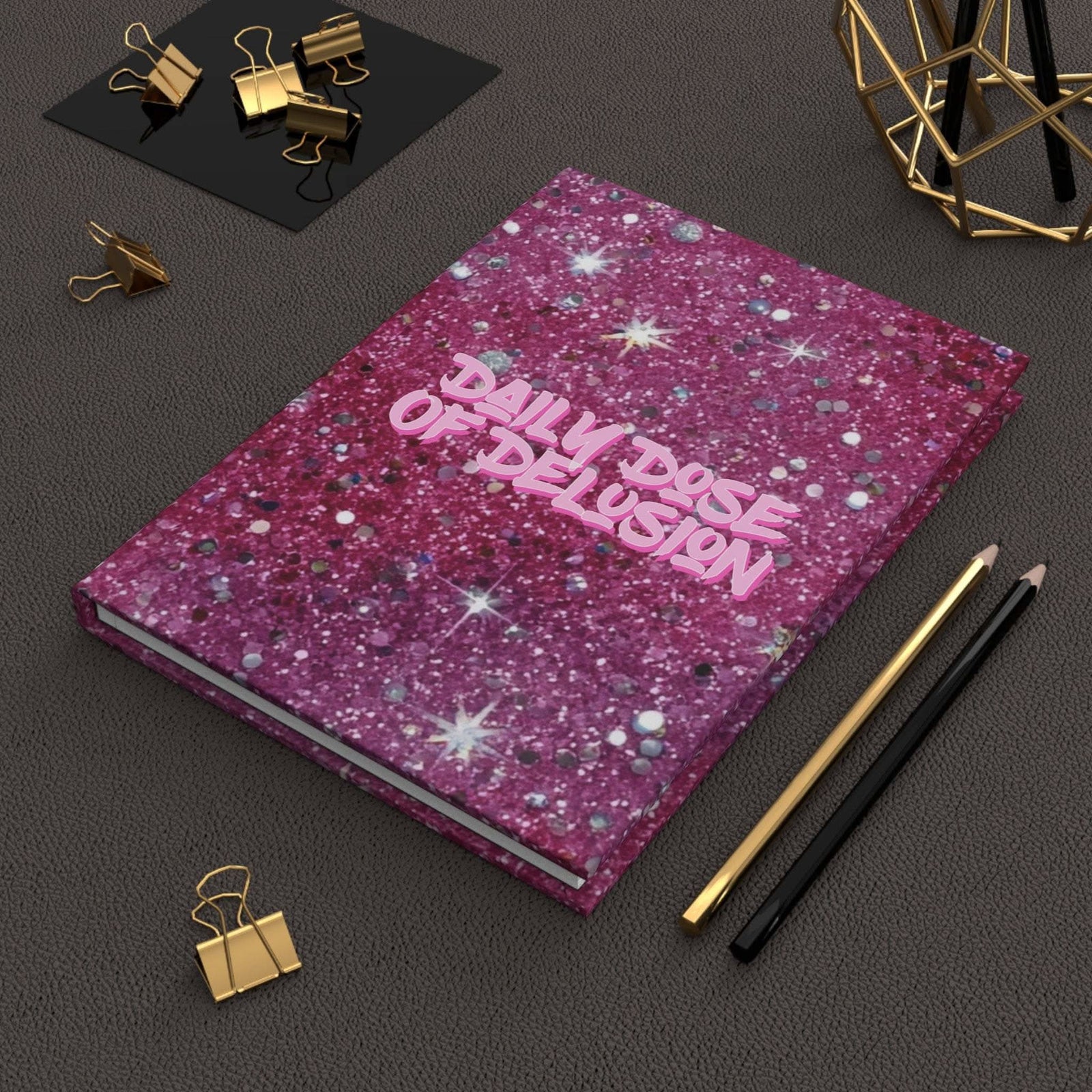 Hardcover journal with pink glitter cover titled Daily Dose of Delusion, on a dark textured surface with pencils and gold binder clips.