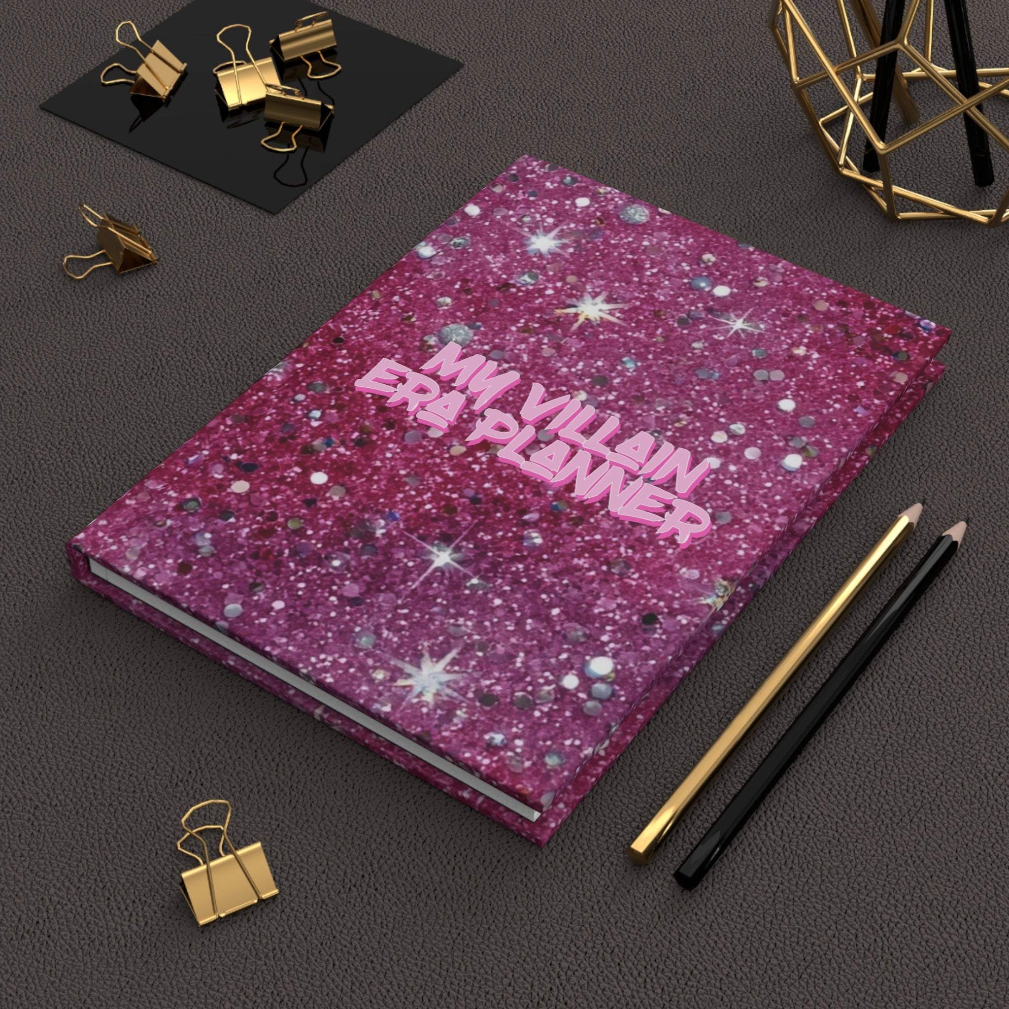 Matte hardcover journal with vibrant pink glitter design titled My Villain Era Planner on black desk with gold and black stationery accessories