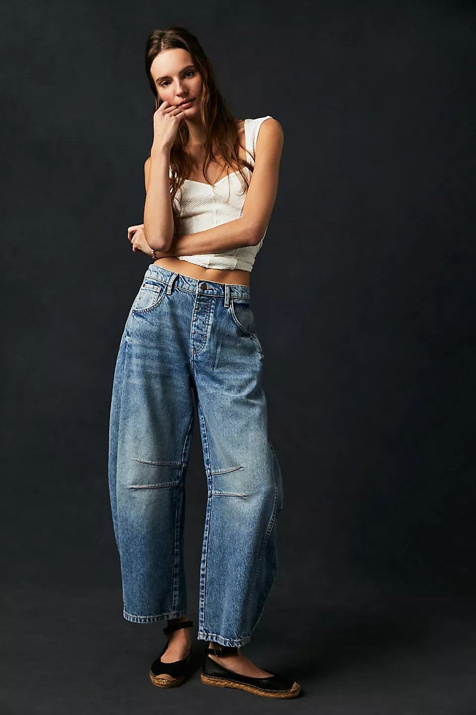 Woman wearing high stretch mid-rise wide leg loose fit denim jeans and white tank top against dark background