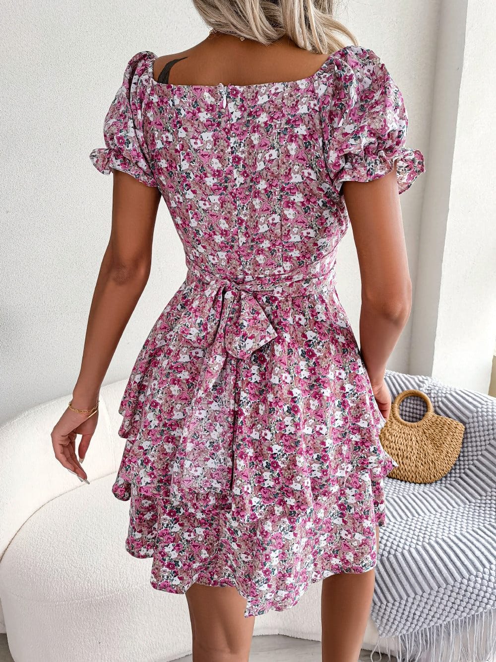 Back view of a women mini dress with pink floral print, puff short sleeves, ruffled hem, and tied waist belt.
