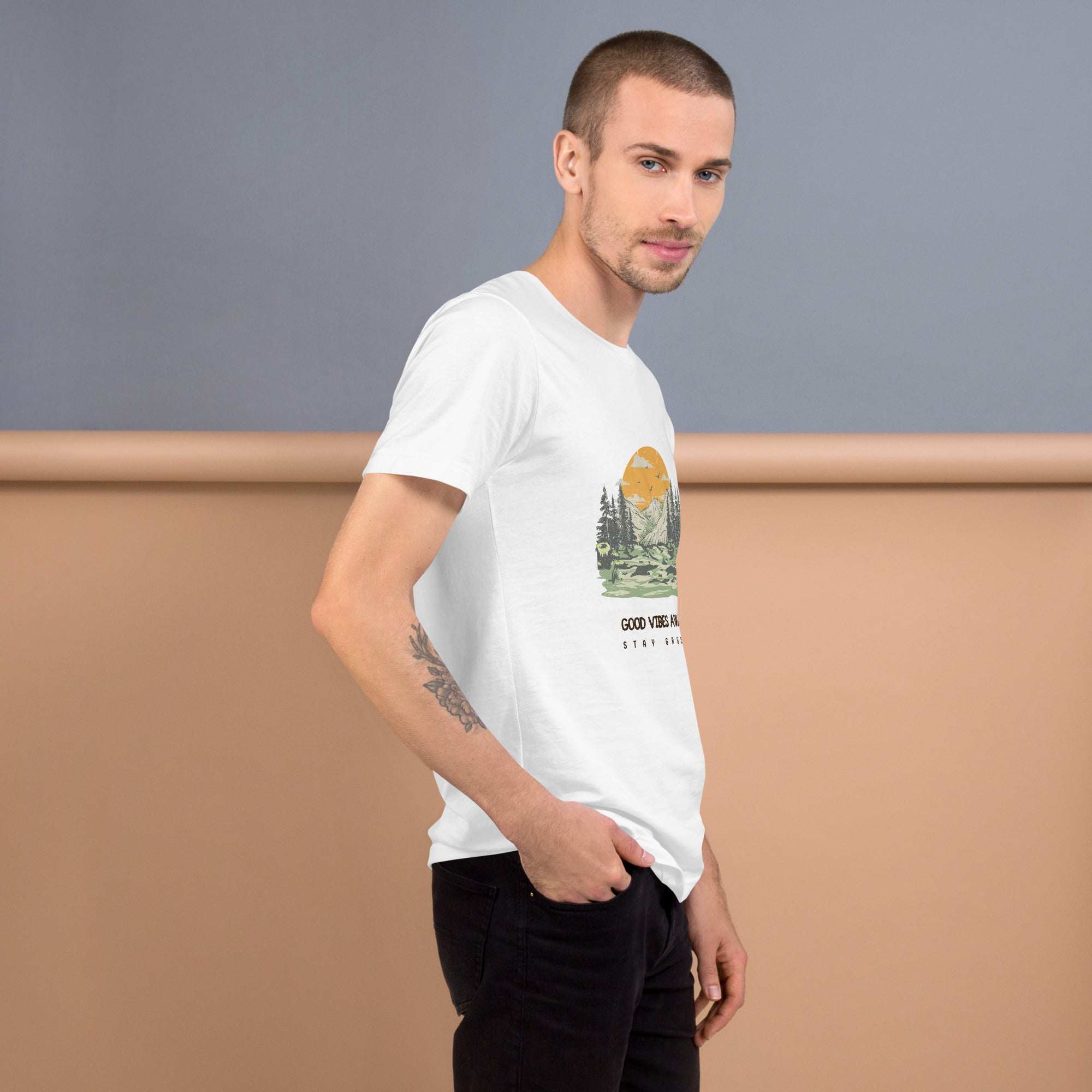 Man wearing white Good Vibes T-Shirt with nature graphic and text on front, posing against gray and beige background.