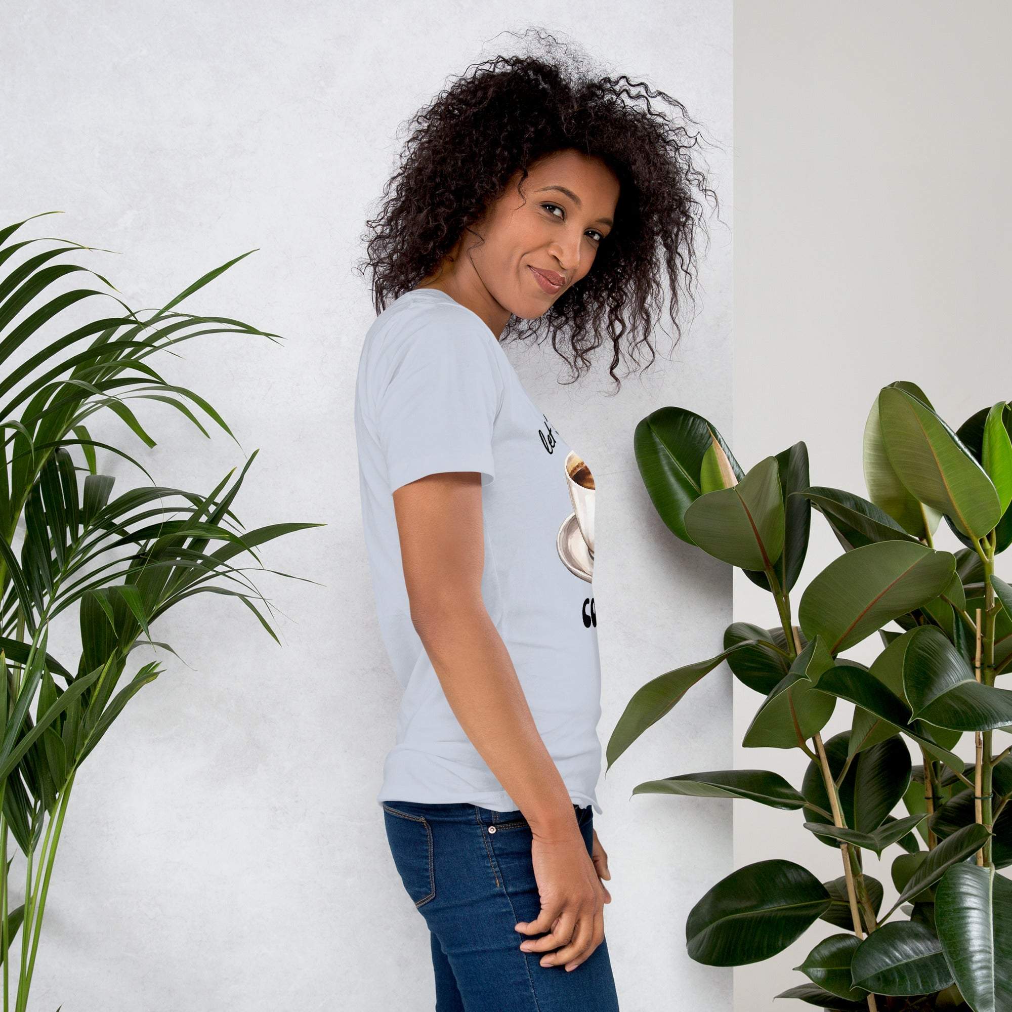 Woman wearing a light blue coffee t-shirt with "Let's Have Some Coffee" design standing near green plants
