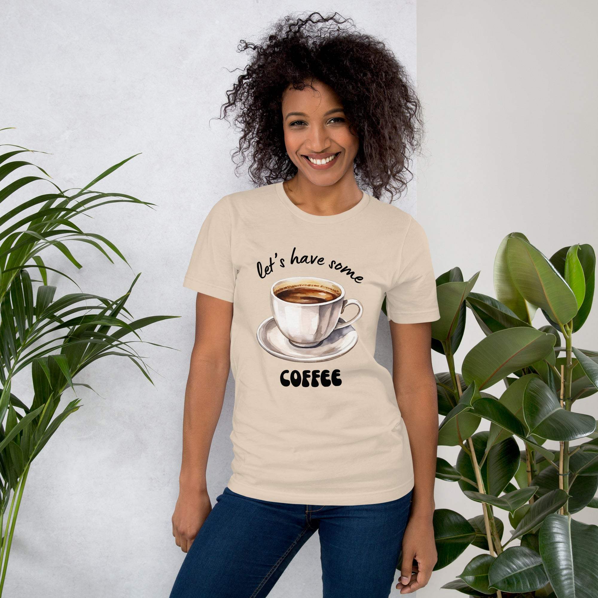 Woman wearing beige "Let's Have Some Coffee" t-shirt with coffee cup graphic and text, smiling indoors near green plants