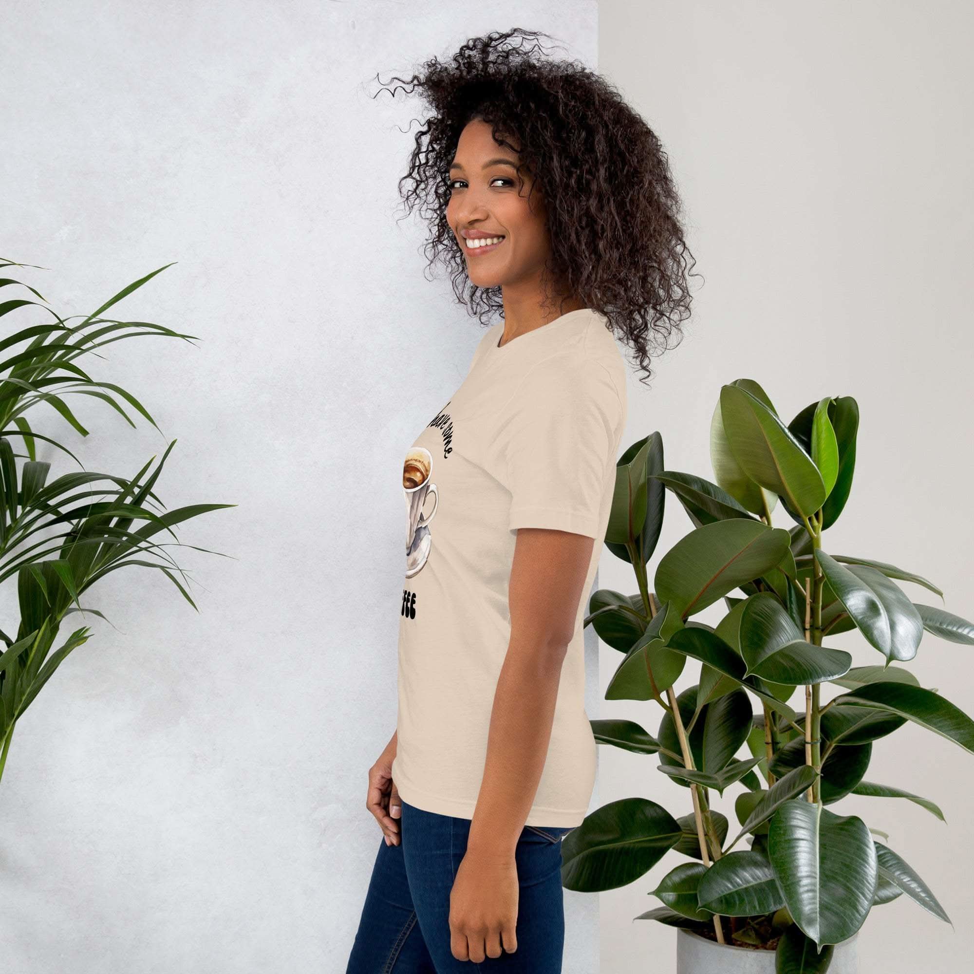 Woman wearing beige Let's Have Some Coffee t-shirt standing next to green plants against white wall background