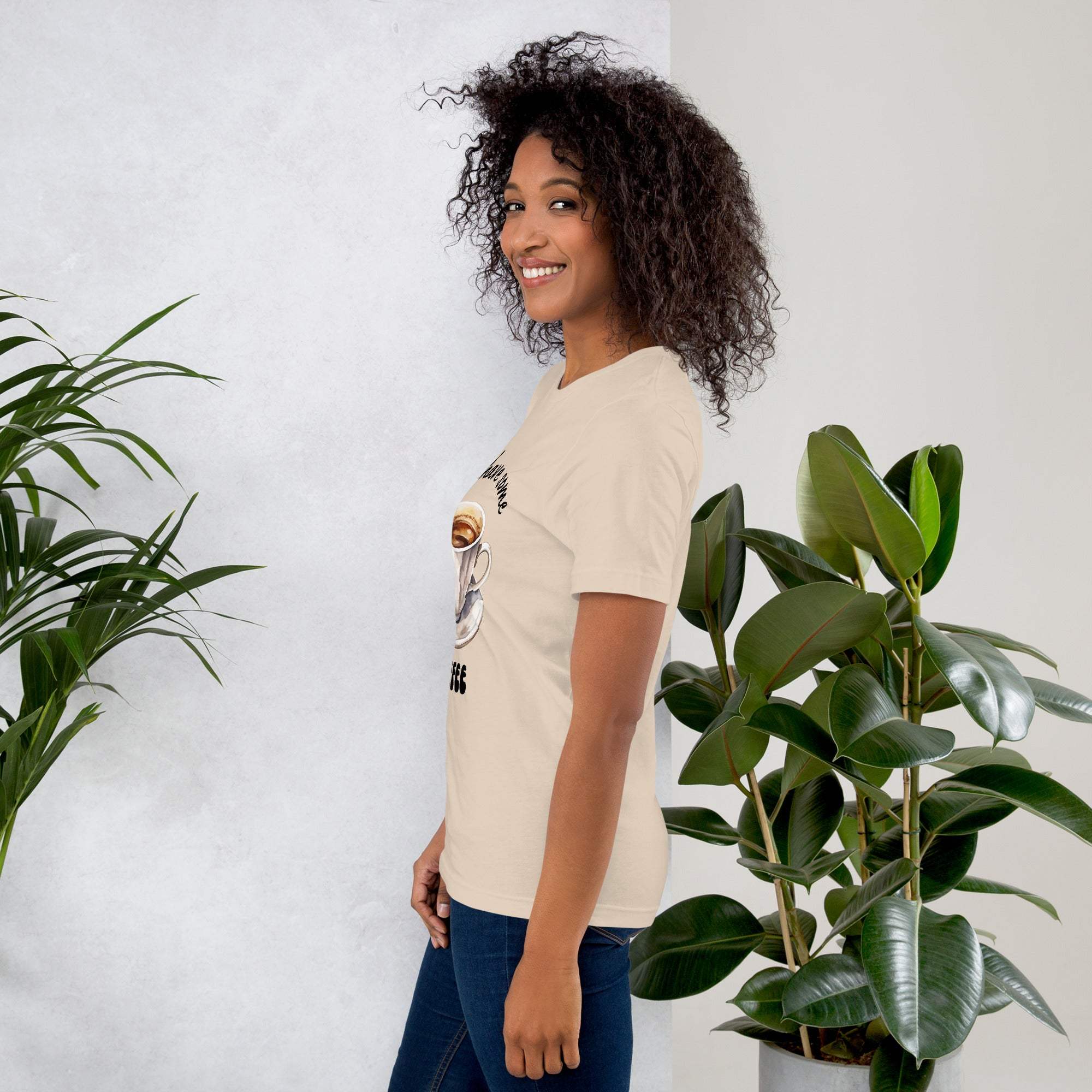 Woman wearing beige Let's Have Some Coffee t-shirt standing next to green plants against white wall background