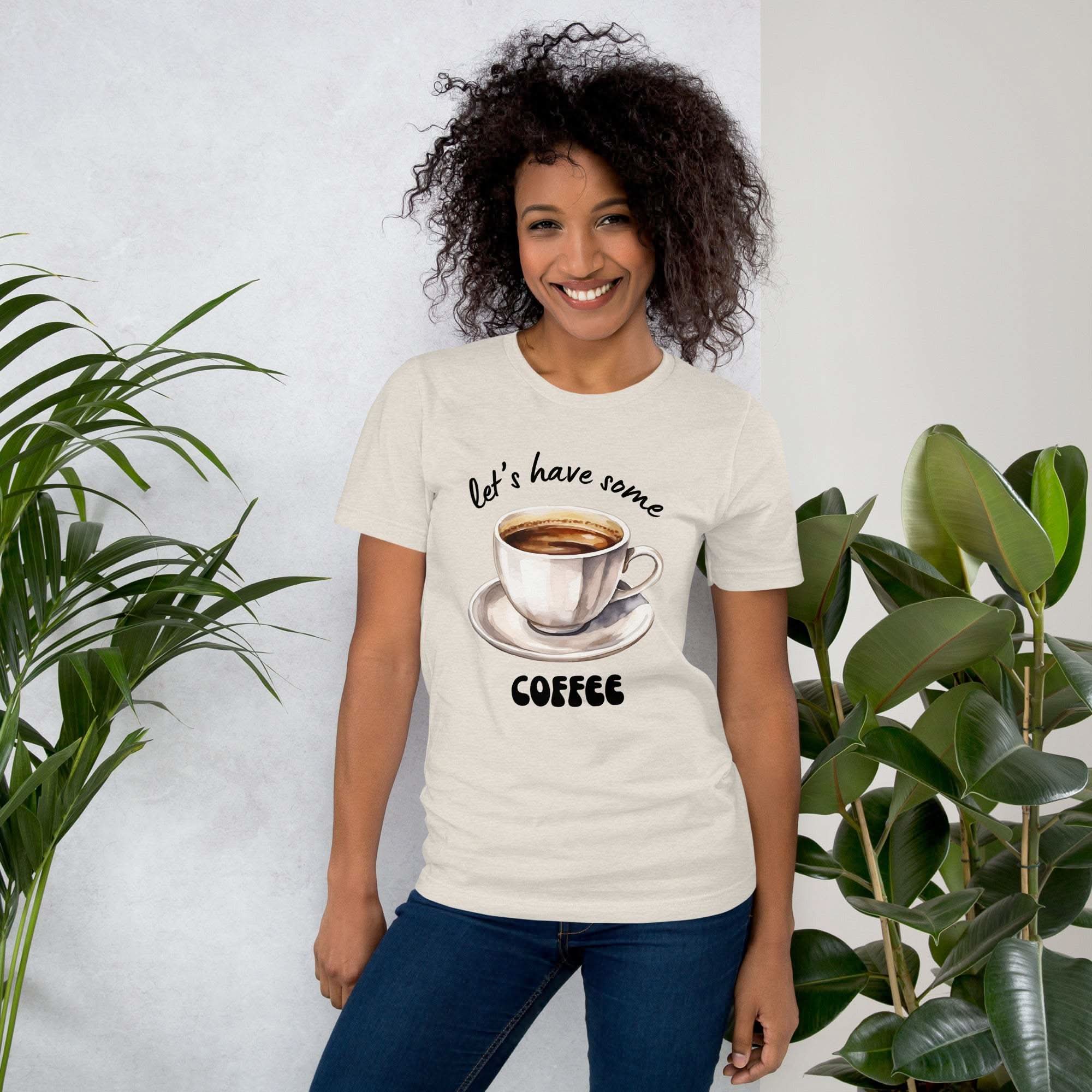 Woman wearing a beige "Let's Have Some Coffee" t-shirt with coffee cup graphic, standing indoors with plants.