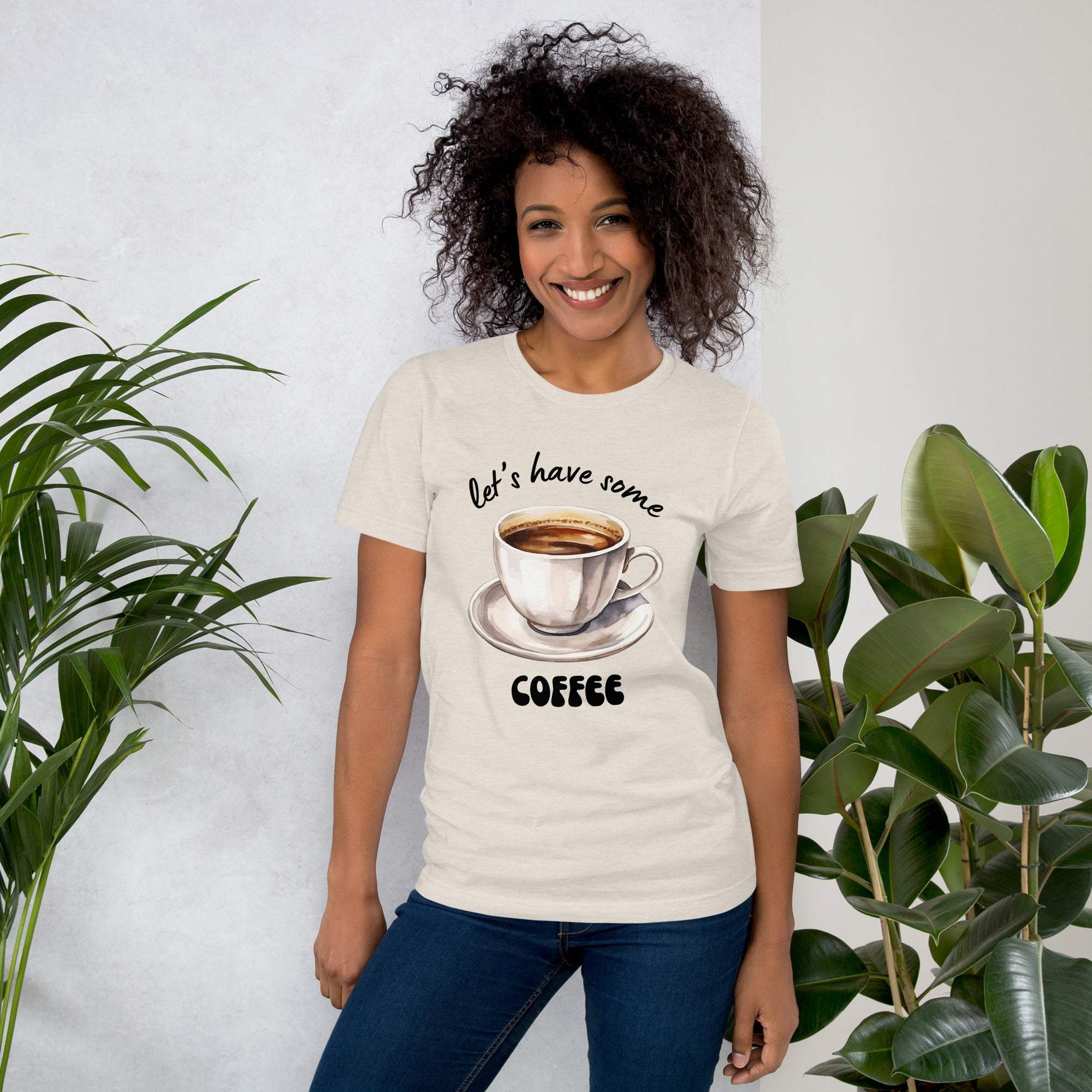 Woman wearing a beige "Let's Have Some Coffee" t-shirt with coffee cup graphic, standing indoors with plants.