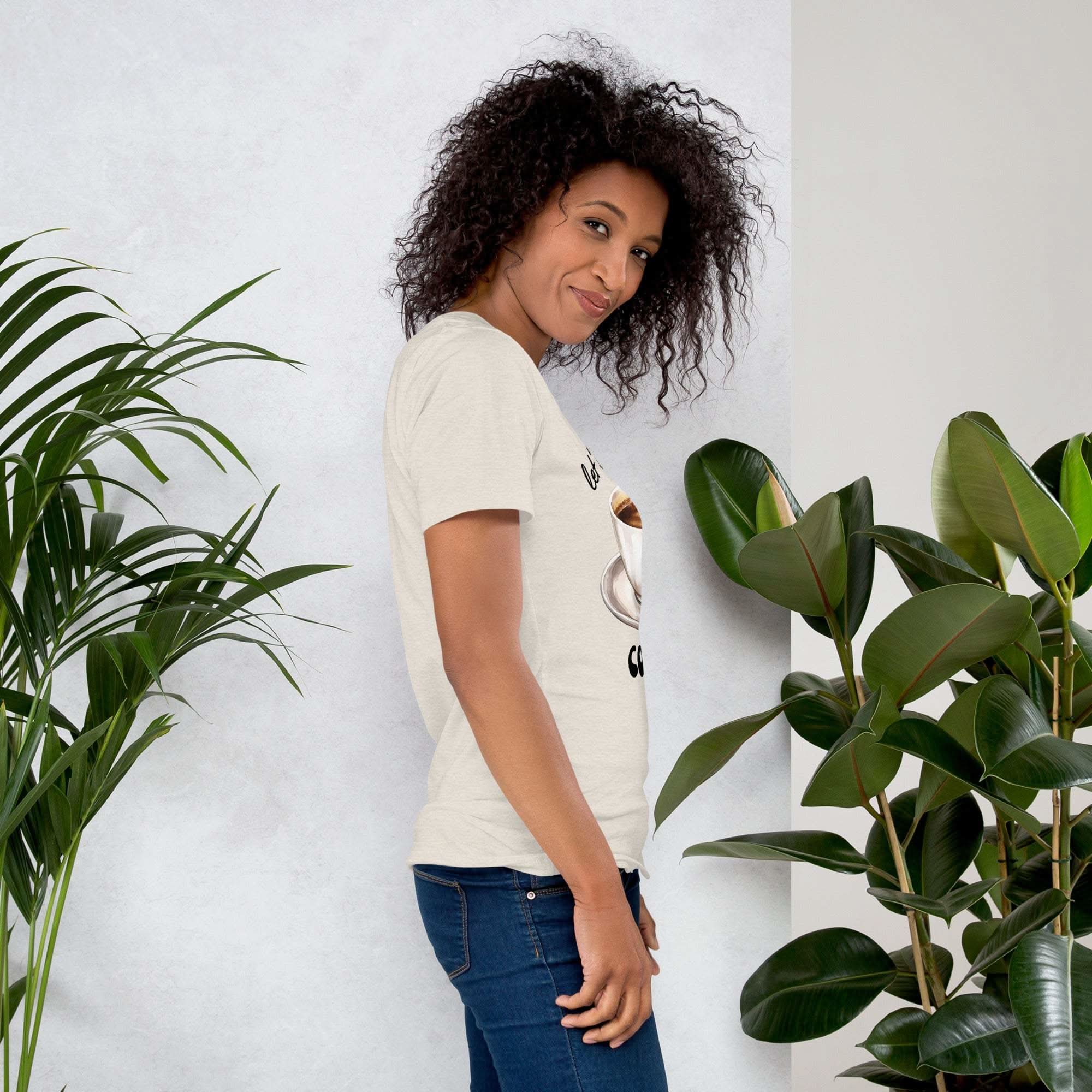 Woman wearing a beige coffee-themed t-shirt with "Let's Have Some Coffee" text standing beside green plants