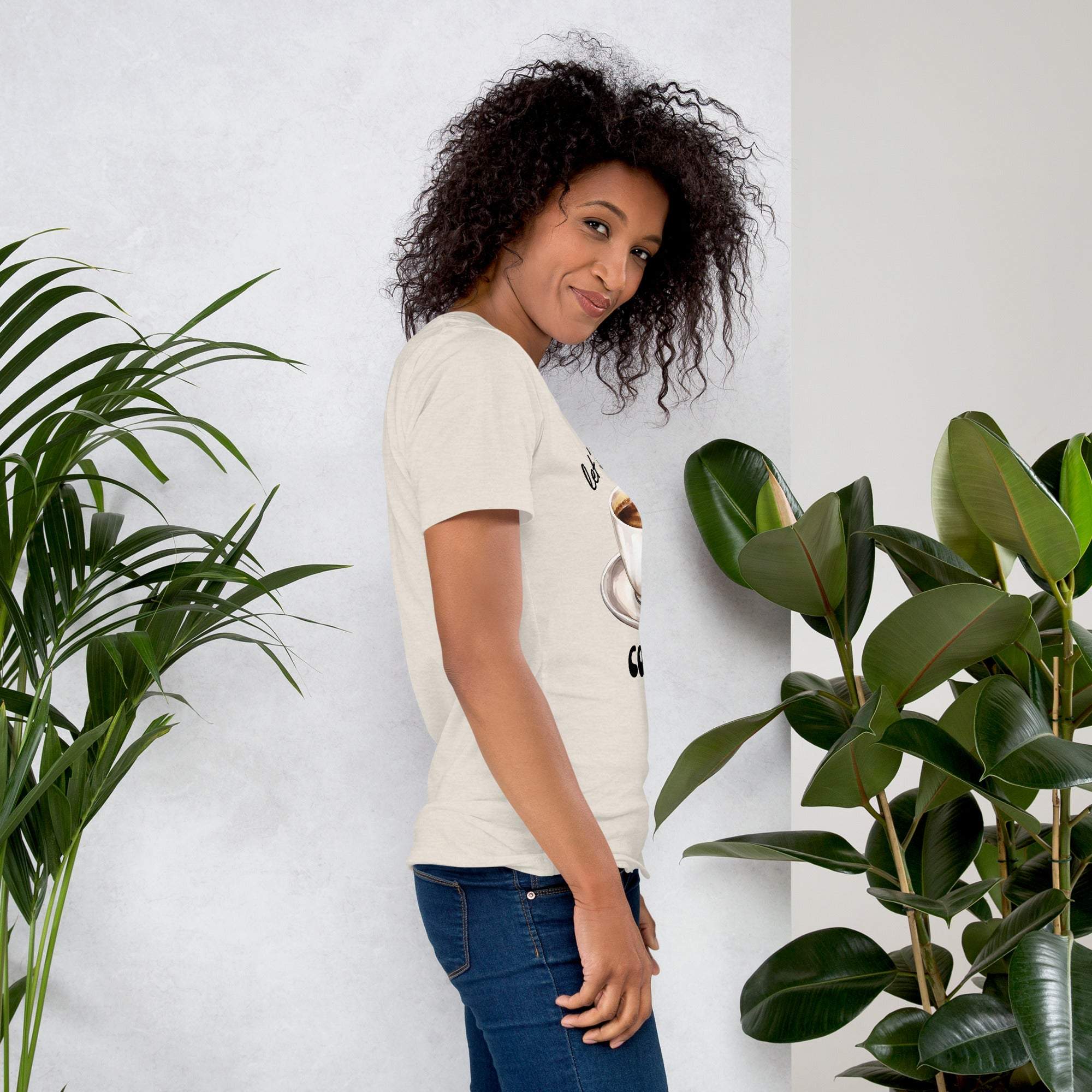 Woman wearing a beige coffee-themed t-shirt with "Let's Have Some Coffee" text standing beside green plants