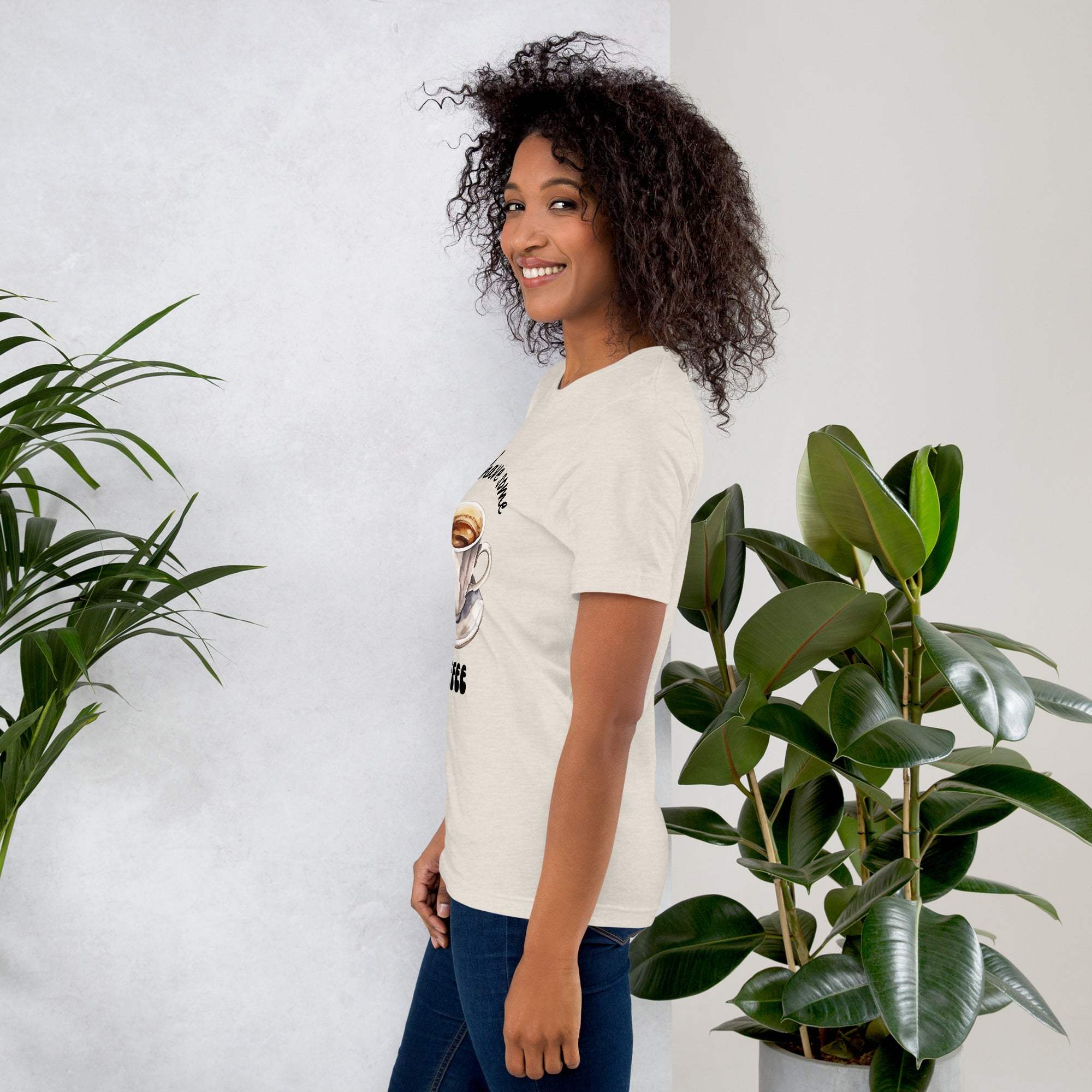 Woman wearing a beige Coffee T-Shirt with a coffee-themed design standing next to green plants