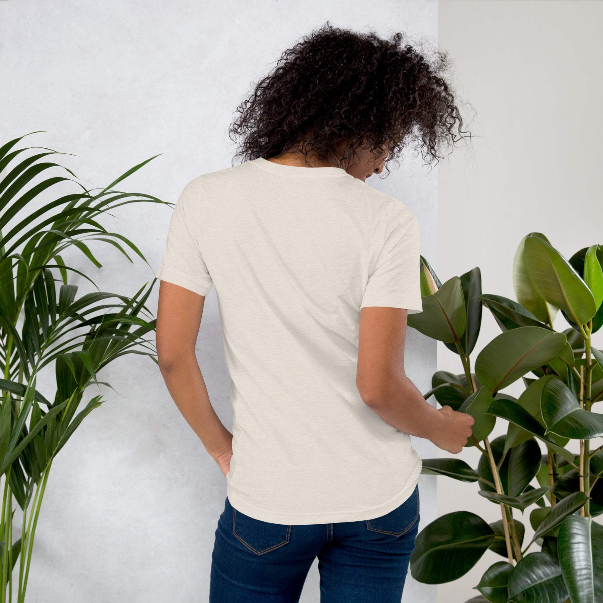 Back view of a woman wearing a beige coffee-themed t-shirt surrounded by green plants