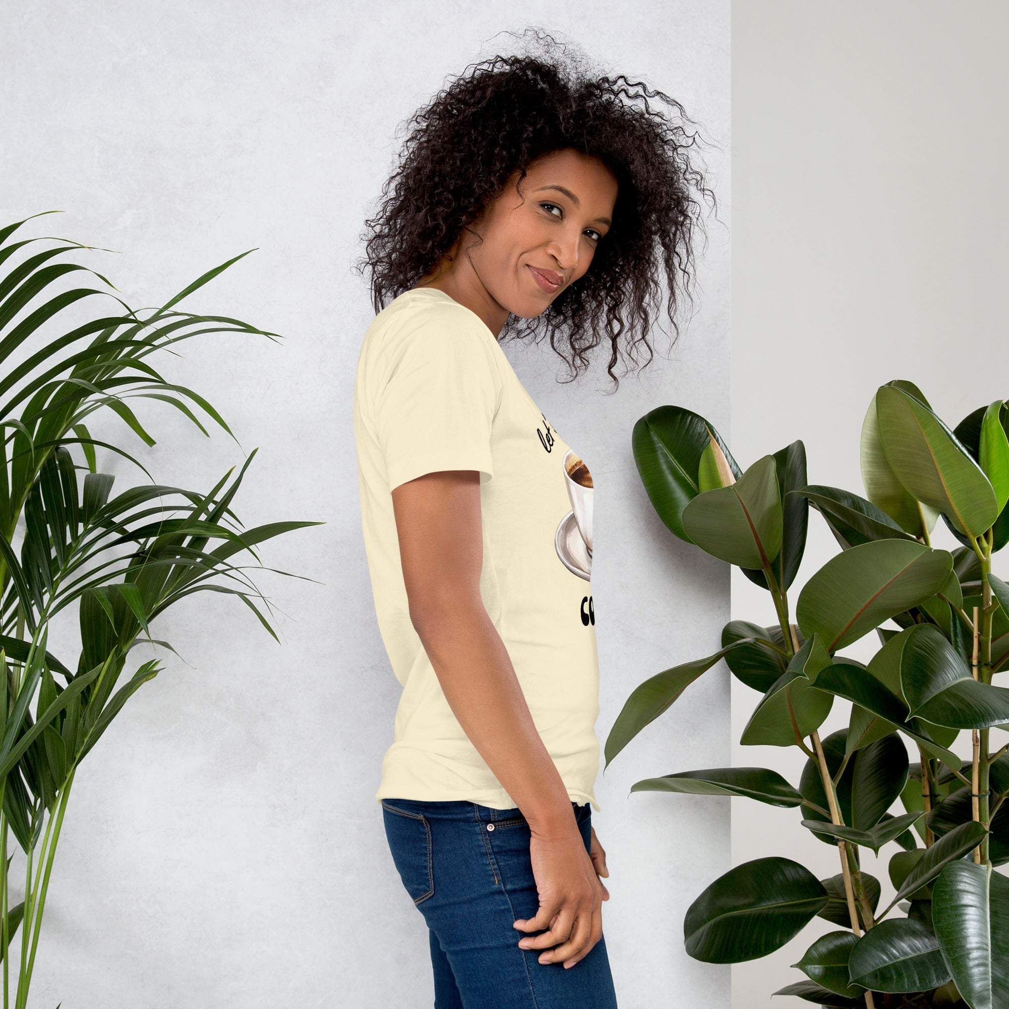 Woman wearing beige coffee t-shirt with coffee cup design standing next to green plants