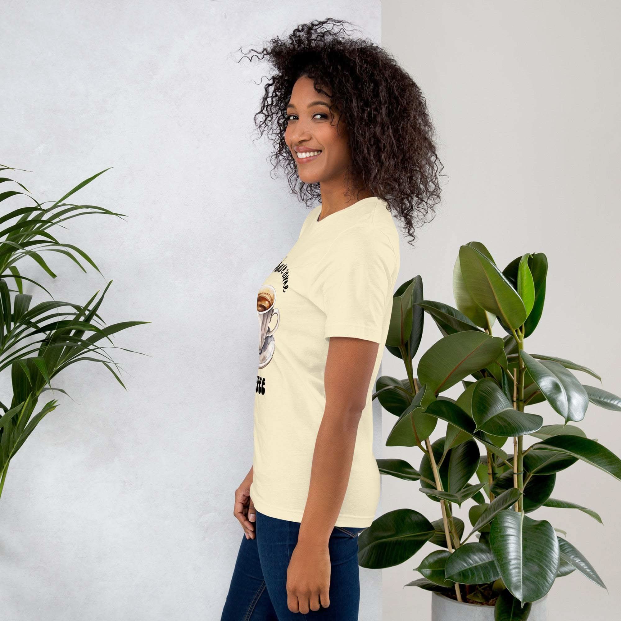 Woman wearing cream coffee t-shirt with "Let's Have Some Coffee" design, standing next to green plants