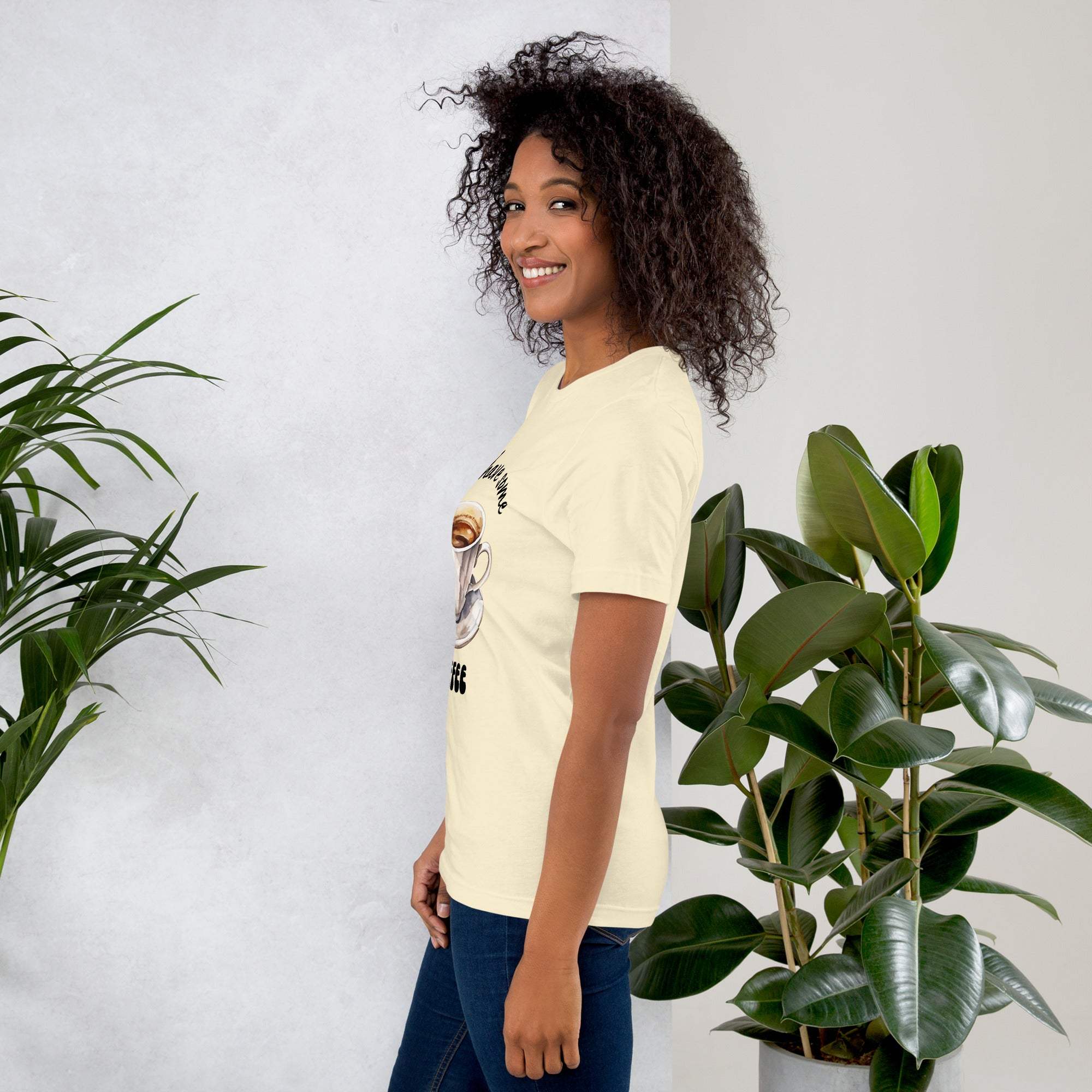 Woman wearing cream coffee t-shirt with "Let's Have Some Coffee" design, standing next to green plants