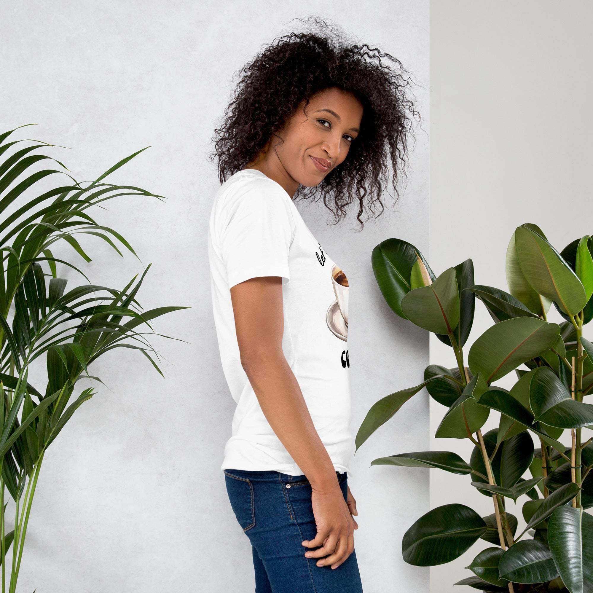Woman wearing white Let's Have Some Coffee coffee t-shirt standing near green plants