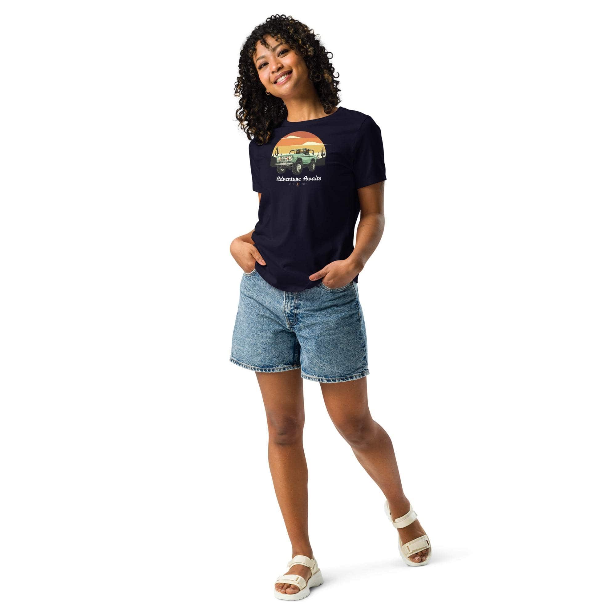 Woman wearing navy Adventure Awaits T-Shirt with off-road vehicle and desert sunset design paired with denim shorts