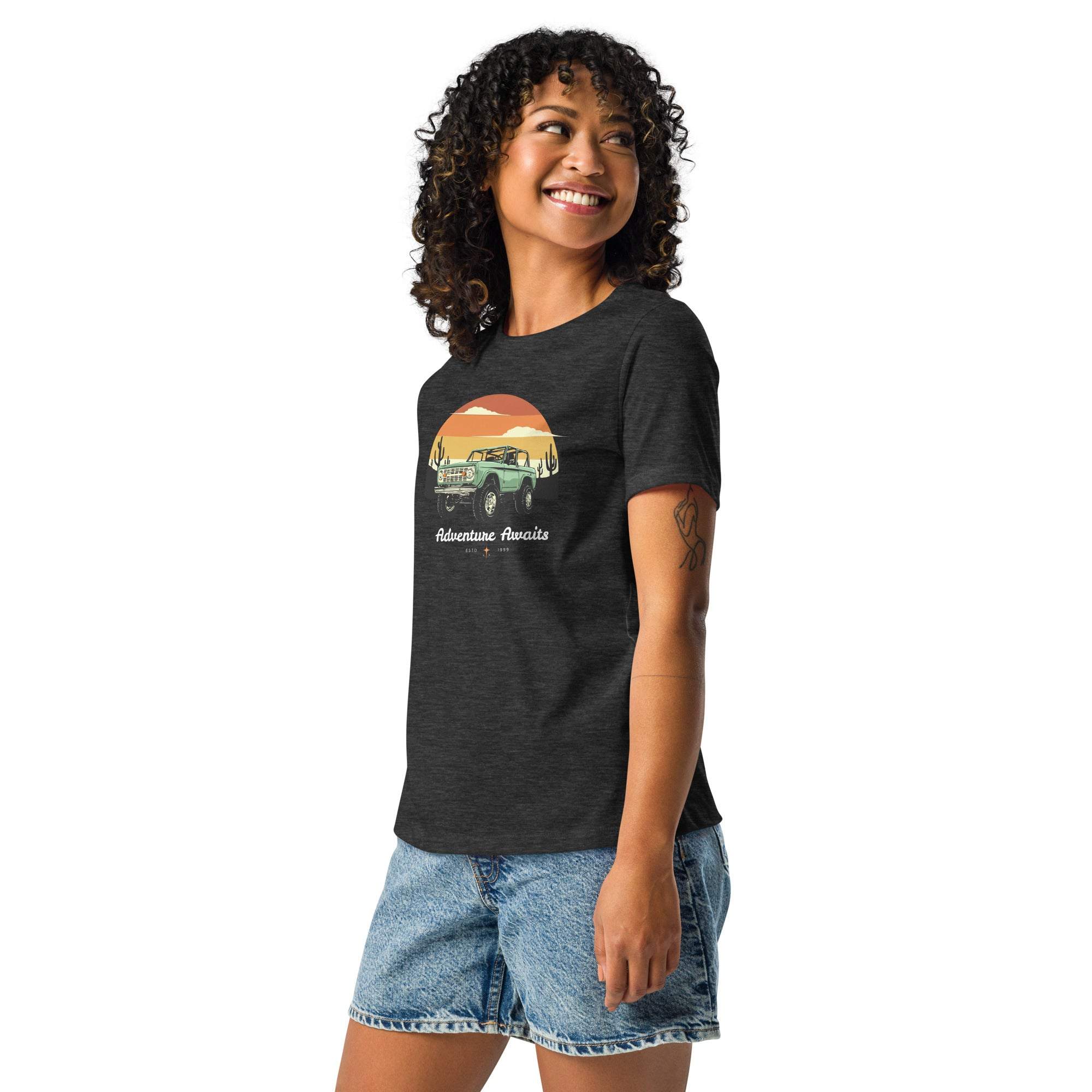 Woman wearing black Adventure Awaits T-Shirt with off-road vehicle and desert sunset design paired with denim shorts