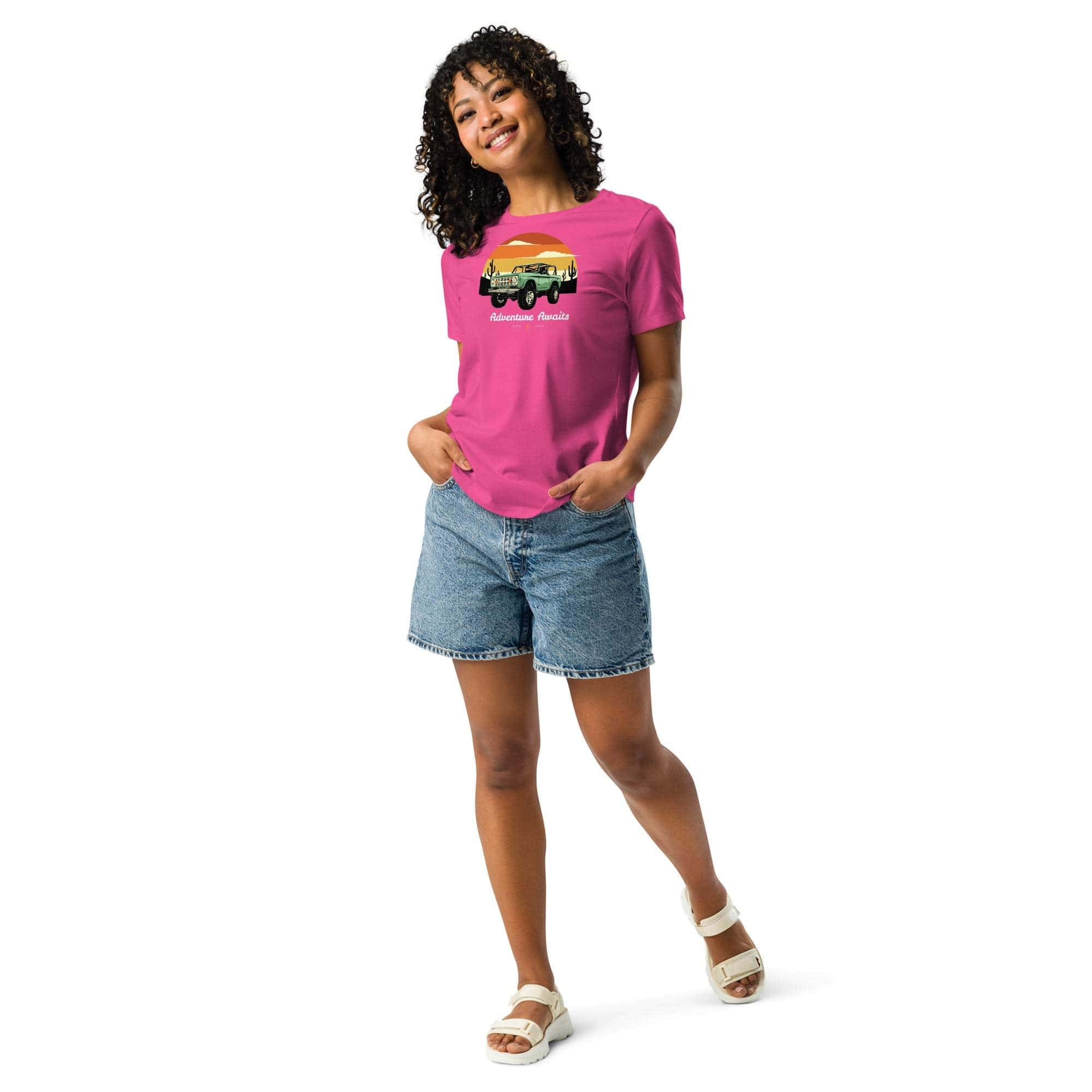 Woman wearing pink Adventure Awaits T-Shirt with off-road vehicle and desert sunset design paired with denim shorts